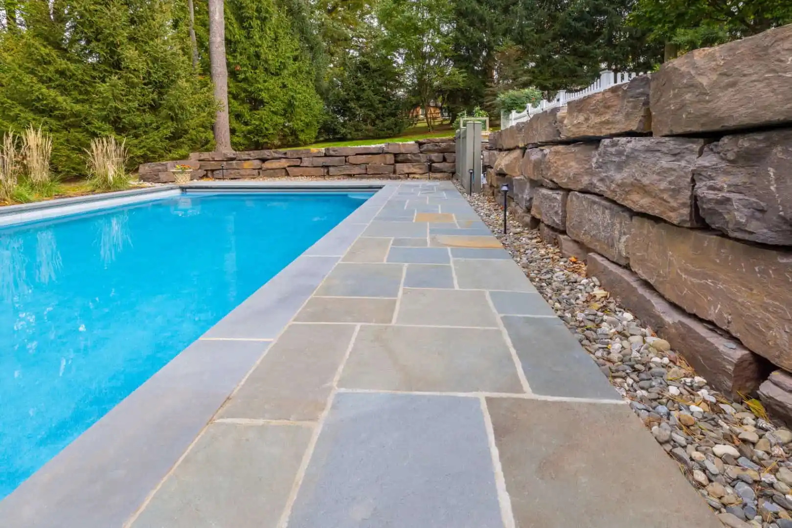 A swimming pool with clear blue water is surrounded by a patio of large stone tiles. To the right, there's a rock wall and decorative gravel. Enhance your outdoor living in Delaware County, PA, with expert hardscaping services to bring this vision to life amidst lush trees and greenery.