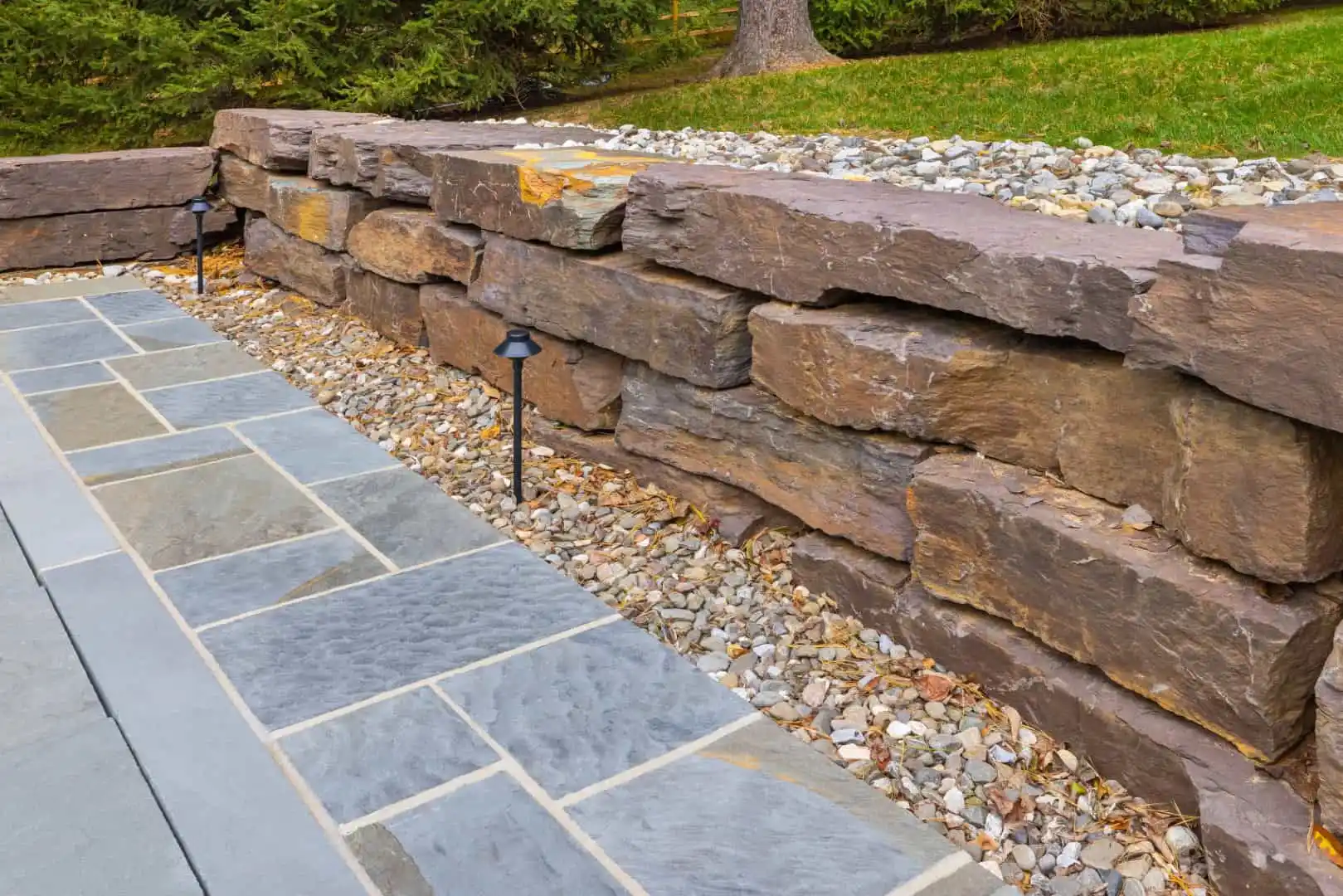 A neatly stacked stone retaining wall, illuminated by two black outdoor lights at its base, is complemented by a paved stone walkway in the foreground. Grass and trees flourish in the background, showcasing the elegance of hardscaping services Delaware County PA offers.