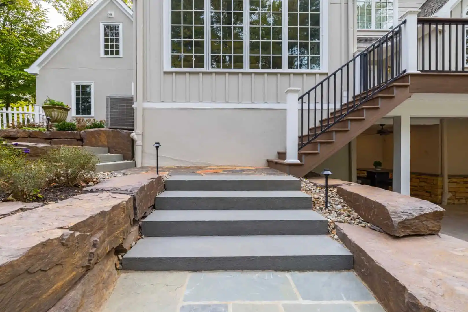 Stone steps lead to a modern house with large windows. A wooden staircase with a railing ascends to the right. Expert landscaping services in Delaware County, PA, have enhanced the aesthetic with rock borders and greenery. The house exterior is pale with contrasting trim and siding.