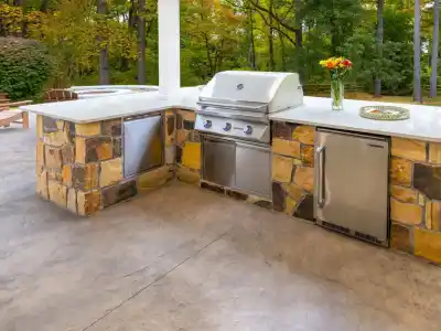 Discover the perfect outdoor living space in Delaware County, PA, with this elegant kitchen. Featuring a stone and stainless steel setup, it includes a gas grill, refrigerator, and shelving. Surrounded by lush greenery, it epitomizes top-tier landscaping services available in the area.
