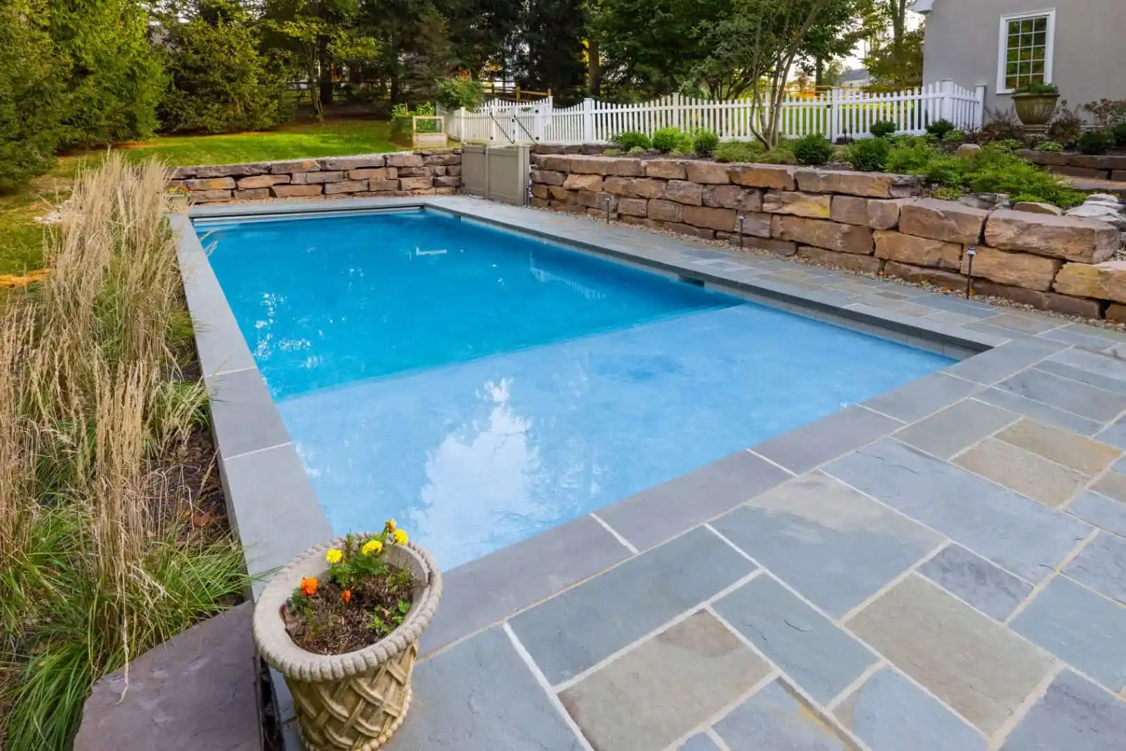 A rectangular swimming pool with clear blue water is surrounded by stone tiles, showcasing expert hardscaping services in Delaware County, PA. A stone retaining wall borders one side, complemented by nearby grass and plants. Bright blooms sit poolside, with a white fence completing the serene outdoor living space.