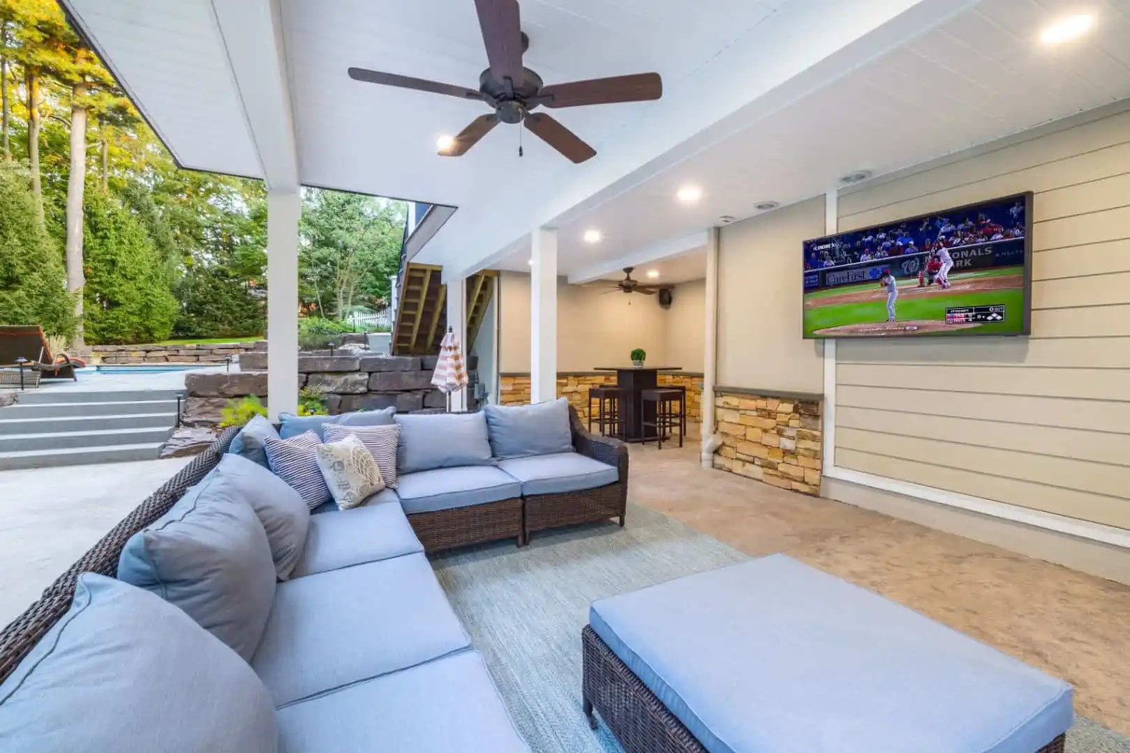 Covered outdoor patio with a ceiling fan, sectional sofa, ottoman, rug, wall-mounted TV showing a baseball game, dining table in the back, and landscaped yard with steps and greenery.