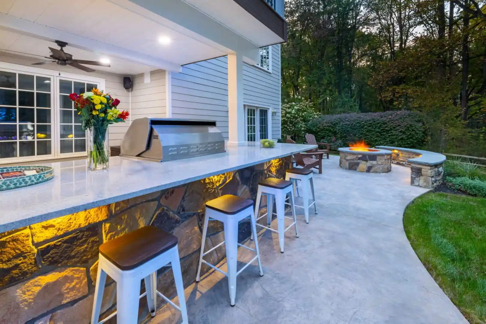 Outdoor living in Delaware County, PA is epitomized by this kitchen and patio with a white stone counter, bar stools, and grill. A vase of flowers and dishes adorn the counter. In the background, there's a fire pit surrounded by chairs amid trees and a white house.