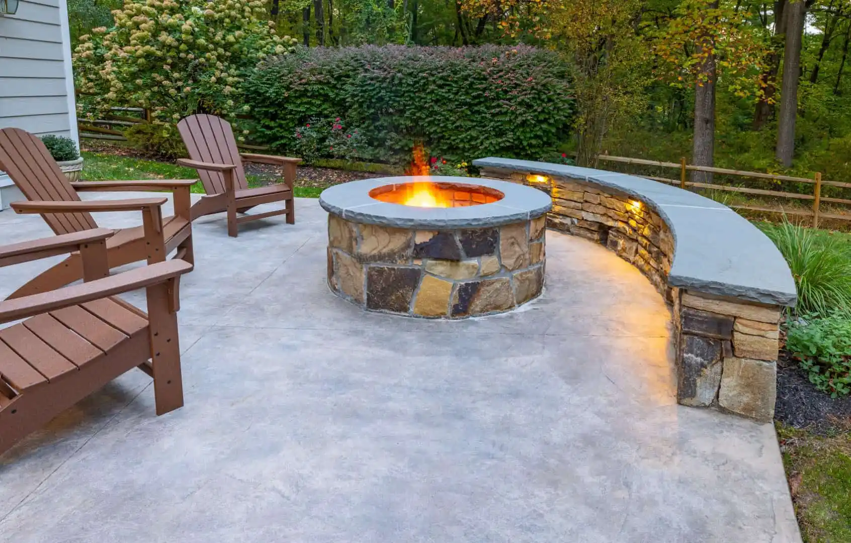 A cozy outdoor patio with a stone fire pit in the center is surrounded by wooden chairs and a curved stone bench with built-in lighting. The area, crafted through expert hardscaping services in Delaware County PA, is bordered by lush greenery and trees.