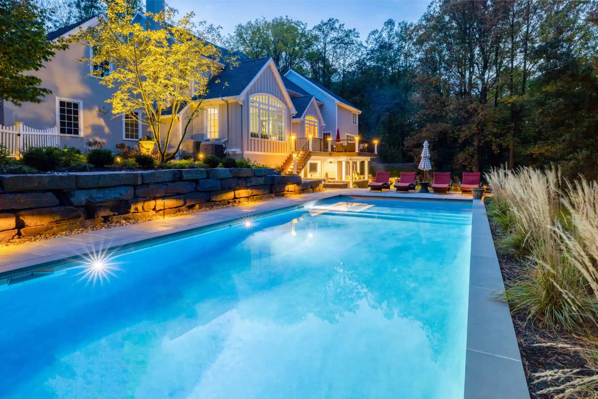 A large, luxurious house with multiple gables is warmly lit at dusk, showcasing outdoor living in Delaware County, PA. It overlooks a swimming pool framed by lounge chairs, tall grass, and a stone wall. Trees and a dimly lit sky enhance this serene haven.