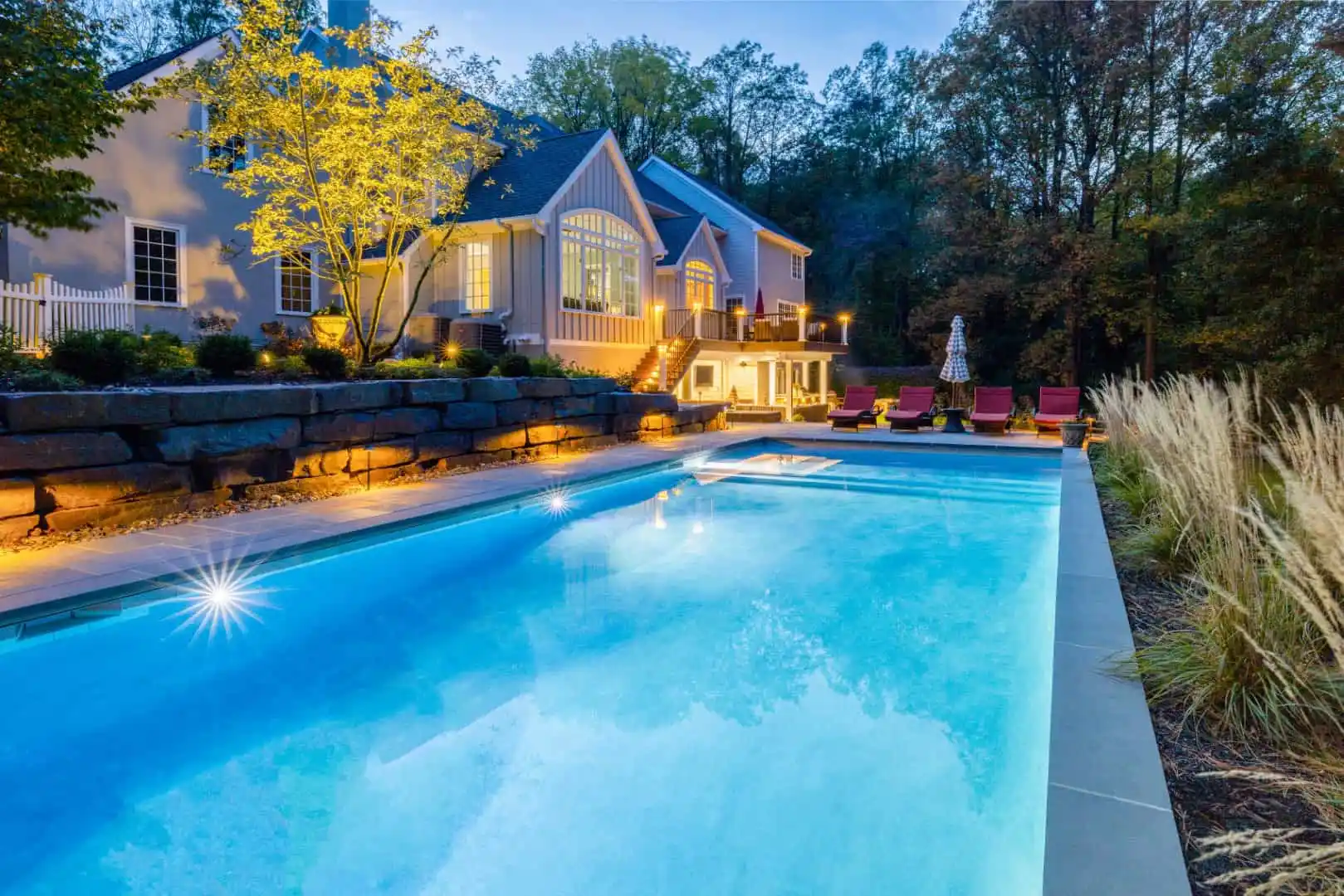 A luxurious house with large windows is illuminated at dusk, overlooking a glowing swimming pool. Surrounded by trees and a stone wall, enhanced by expert landscaping services in Delaware County PA, red lounge chairs are arranged poolside, crafting a serene and elegant outdoor space.