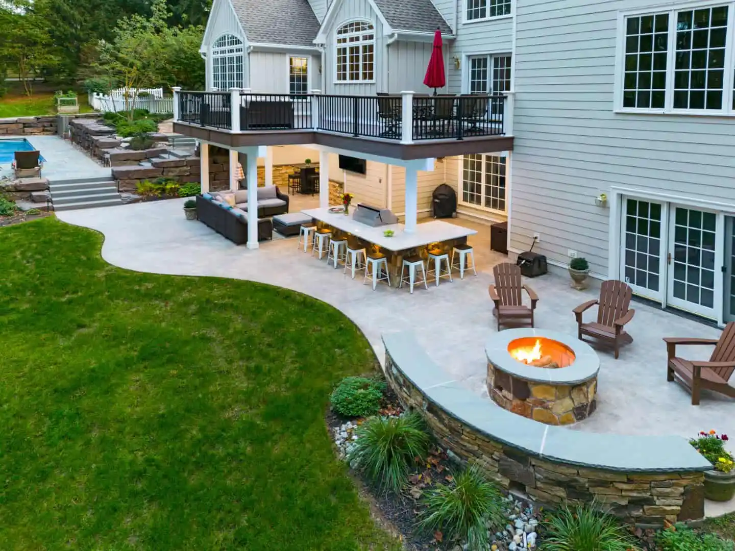 A spacious backyard in Delaware County, PA, features a white house with a large patio area. With exquisite outdoor living elements like cushioned seating and a fire pit surrounded by Adirondack chairs, lush landscaping enhances the green grass while a pool glistens in the background.