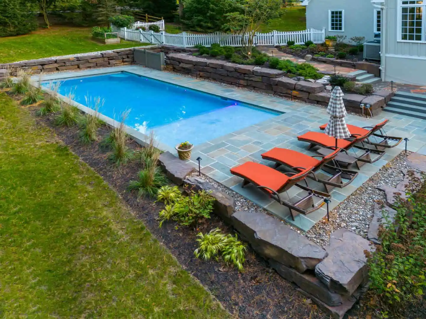 A backyard scene in Delaware County, PA, showcases outdoor living at its finest with a rectangular swimming pool surrounded by stone tiles. Four red lounge chairs and a striped umbrella sit poolside amid lush landscaping. A white picket fence and steps leading to the house complete this serene setting.