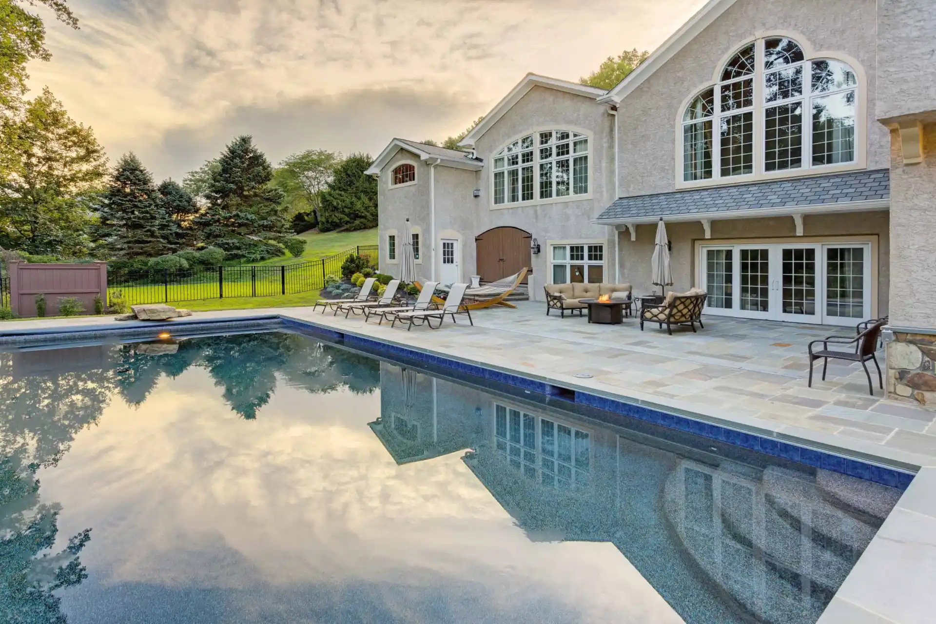 Large modern house with tall arched windows and a spacious stone patio featuring lounge chairs, tables, and umbrellas beside a clear swimming pool reflecting the sky and trees at sunset.