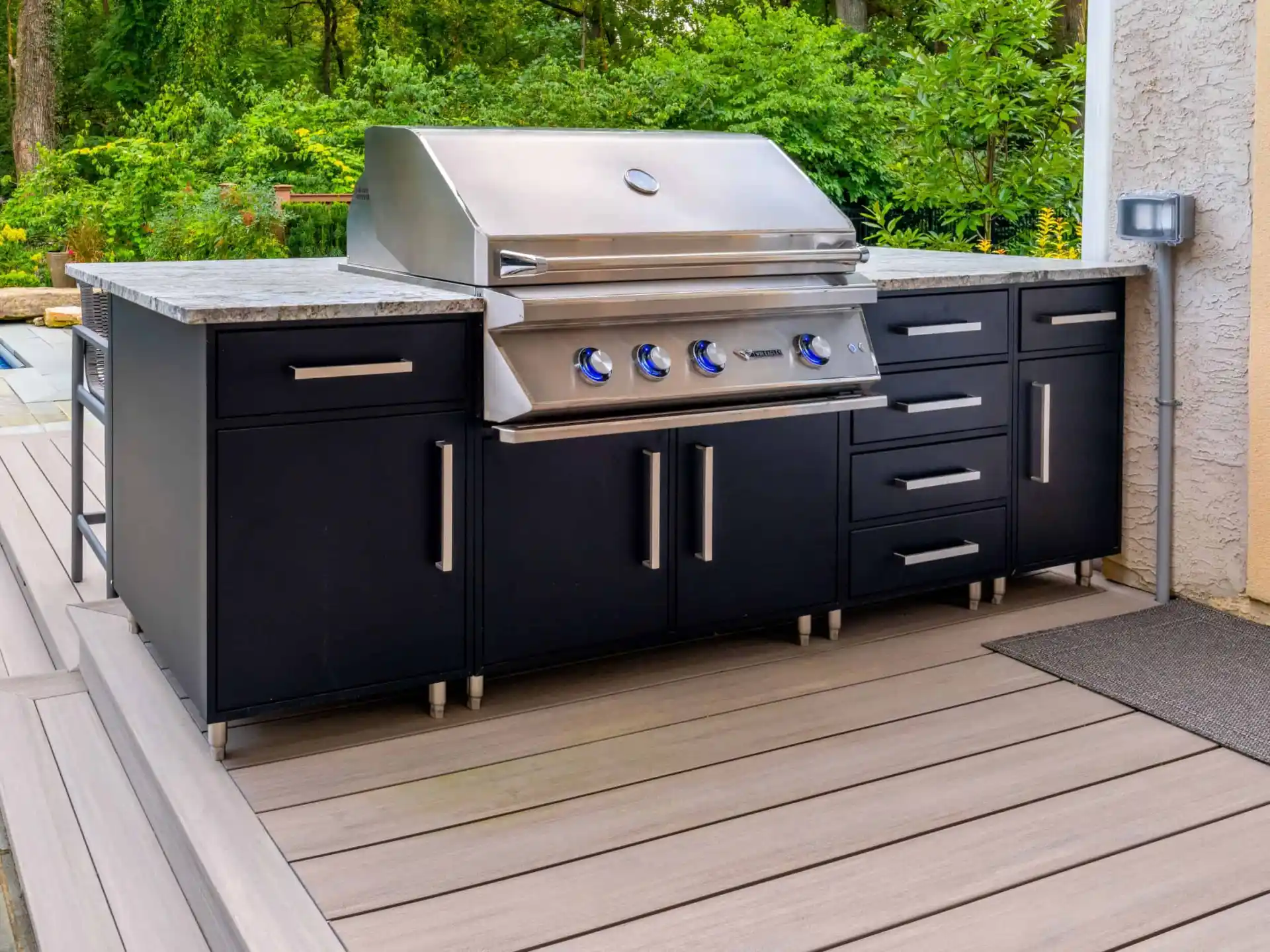 Outdoor living in Delaware County, PA is elevated with this sleek, stainless steel grill on a black cabinet base. Nestled on a wooden deck and surrounded by lush greenery, the grill boasts five control knobs and ample counter space, perfect for any outdoor cooking aficionado.