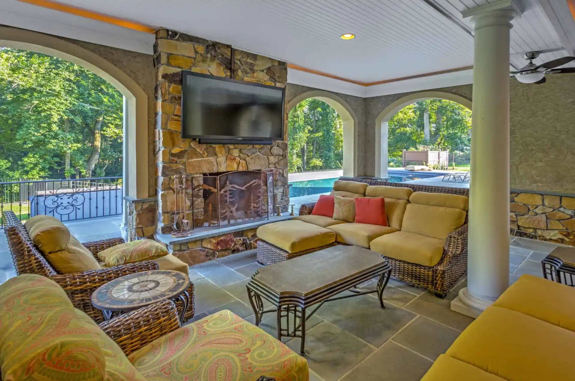 Covered patio with wicker furniture, cushioned seats, and a stone fireplace topped by a mounted TV. Large archways reveal a view of a backyard pool and lush green trees outside.