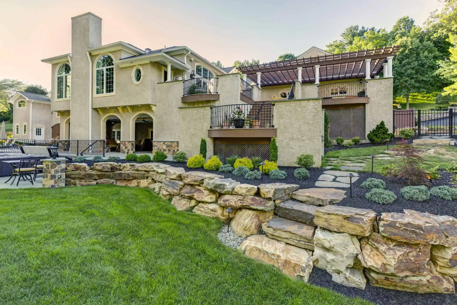 Large two-story beige house with arched windows, multiple balconies, stone accents, a pergola, landscaped garden beds, and a retaining wall made of large rocks, set against a green lawn and trees.