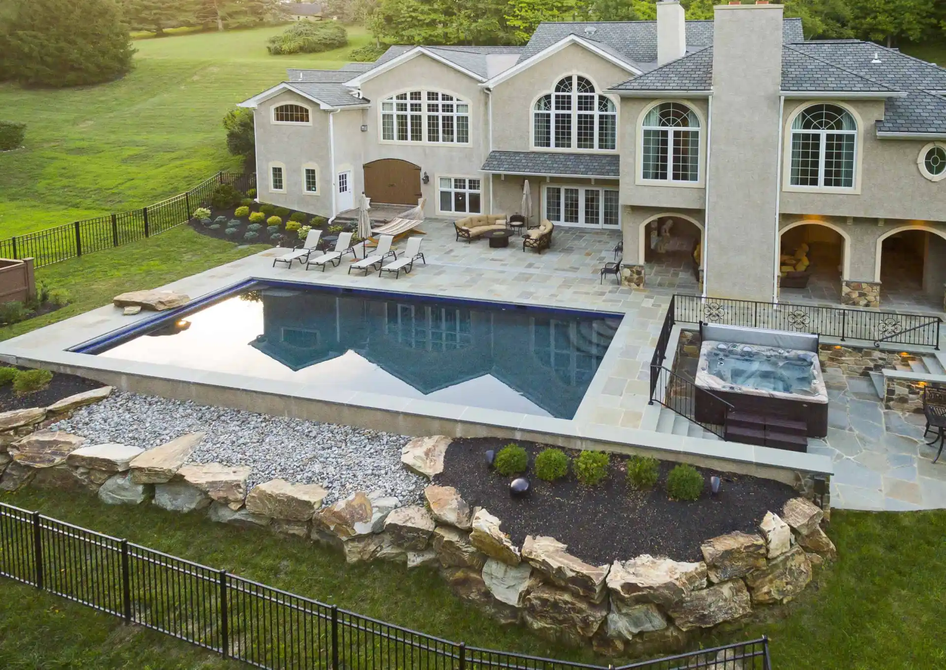 A large, modern house with a spacious backyard featuring a rectangular swimming pool, a hot tub, lounge chairs, stone landscaping, and a patio area, all surrounded by greenery and a black metal fence.