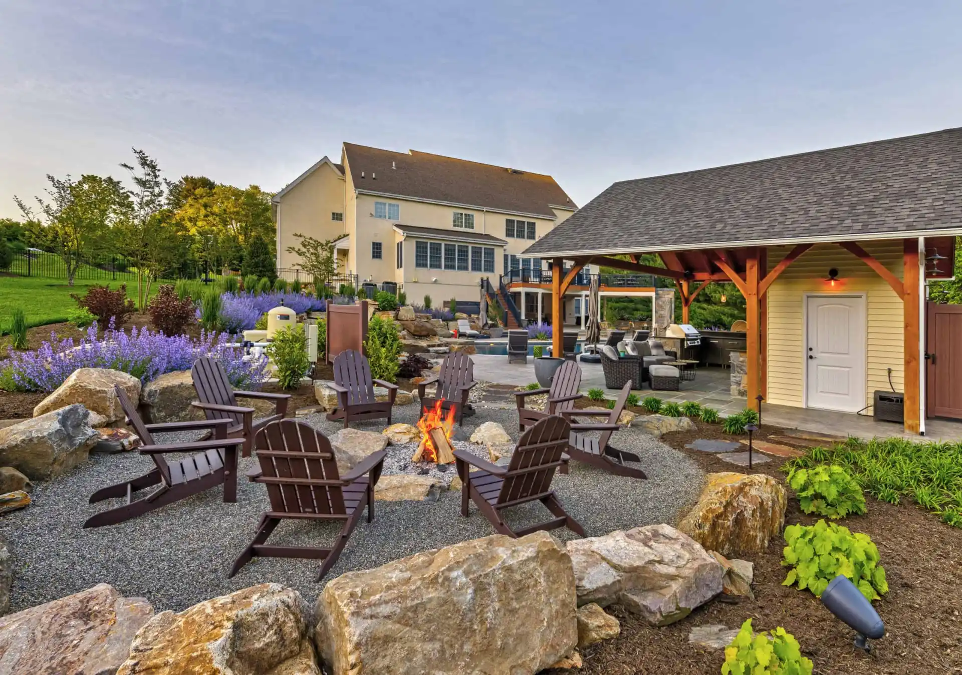 Outdoor seating area with Adirondack chairs around a fire pit, surrounded by rocks and plants. In the background, there’s a large house, a covered patio with furniture, and landscaped gardens under a blue sky.
