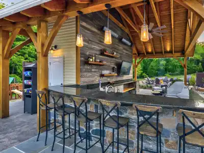 An outdoor bar with a wooden roof and stone counter, surrounded by metal high chairs, embodies the essence of outdoor living in Delaware County PA. Shelves stocked with bottles, a TV, and pendant lights add charm to the space, set against a lush backdrop of trees and an inviting seating area.