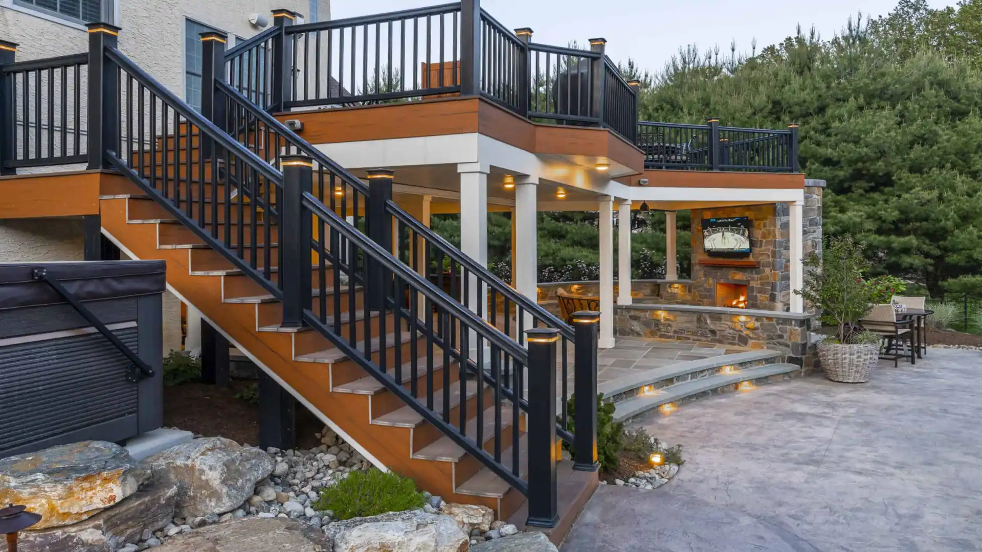 Outdoor living in Delaware County, PA shines with this multi-level patio featuring a staircase to a covered seating area. Enjoy the stone fireplace and elegant lighting fixtures, all expertly designed through our hardscaping services and surrounded by lush greenery and rocks.