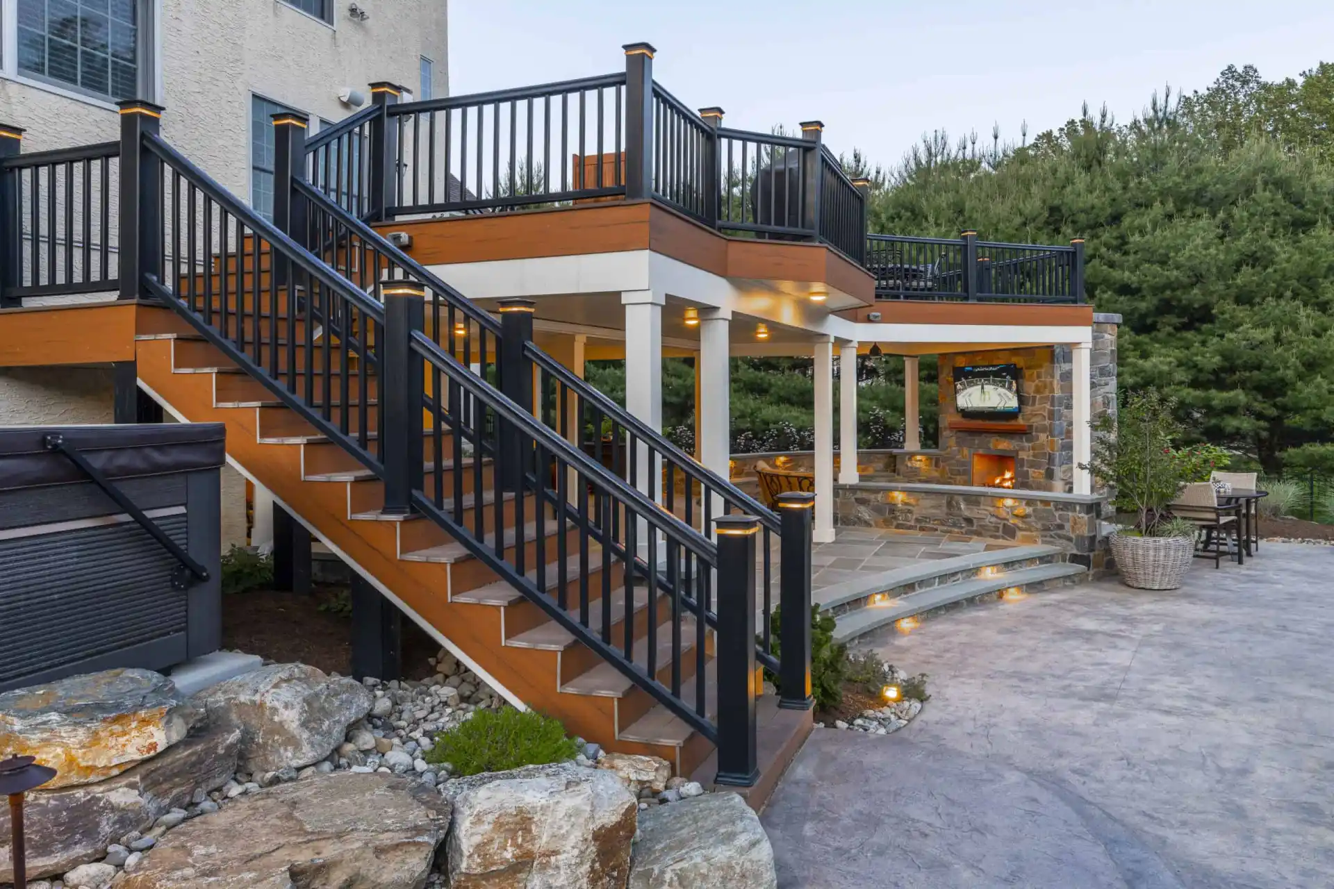 Outdoor living in Delaware County, PA shines with this multi-level patio featuring a staircase to a covered seating area. Enjoy the stone fireplace and elegant lighting fixtures, all expertly designed through our hardscaping services and surrounded by lush greenery and rocks.