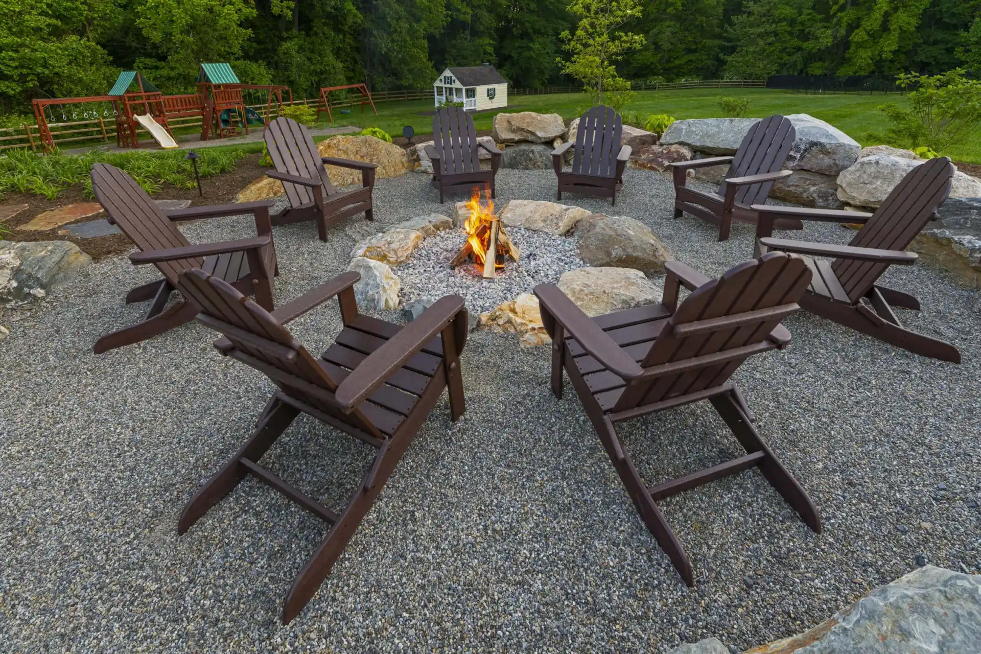 A campfire burns in a stone fire pit surrounded by eight wooden Adirondack chairs on gravel, with a playground and small white shed visible in the grassy background.