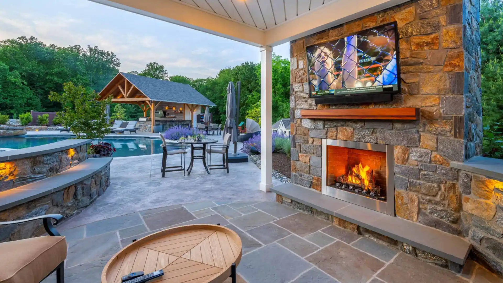 A cozy outdoor patio, crafted with expert hardscaping services in Delaware County, PA, features a stone fire pit with a bright flame. Surrounding the pit are turquoise Adirondack chairs. In the background, stone steps with glowing lights lead upward, flanked by plants and a stone wall.