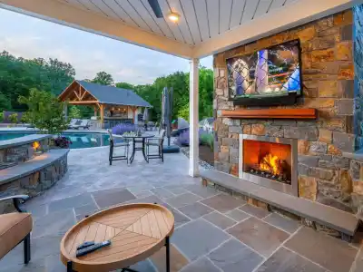 A cozy outdoor patio, crafted with expert hardscaping services in Delaware County, PA, features a stone fire pit with a bright flame. Surrounding the pit are turquoise Adirondack chairs. In the background, stone steps with glowing lights lead upward, flanked by plants and a stone wall.