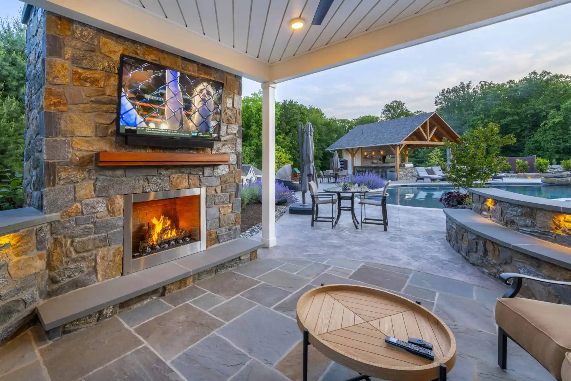 Covered patio with stone fireplace, mounted TV, and burning fire. Outdoor seating, table, and pool area with lounge chairs and a wooden pavilion are visible. Surrounded by trees and lush landscaping. Remote on round coffee table.
