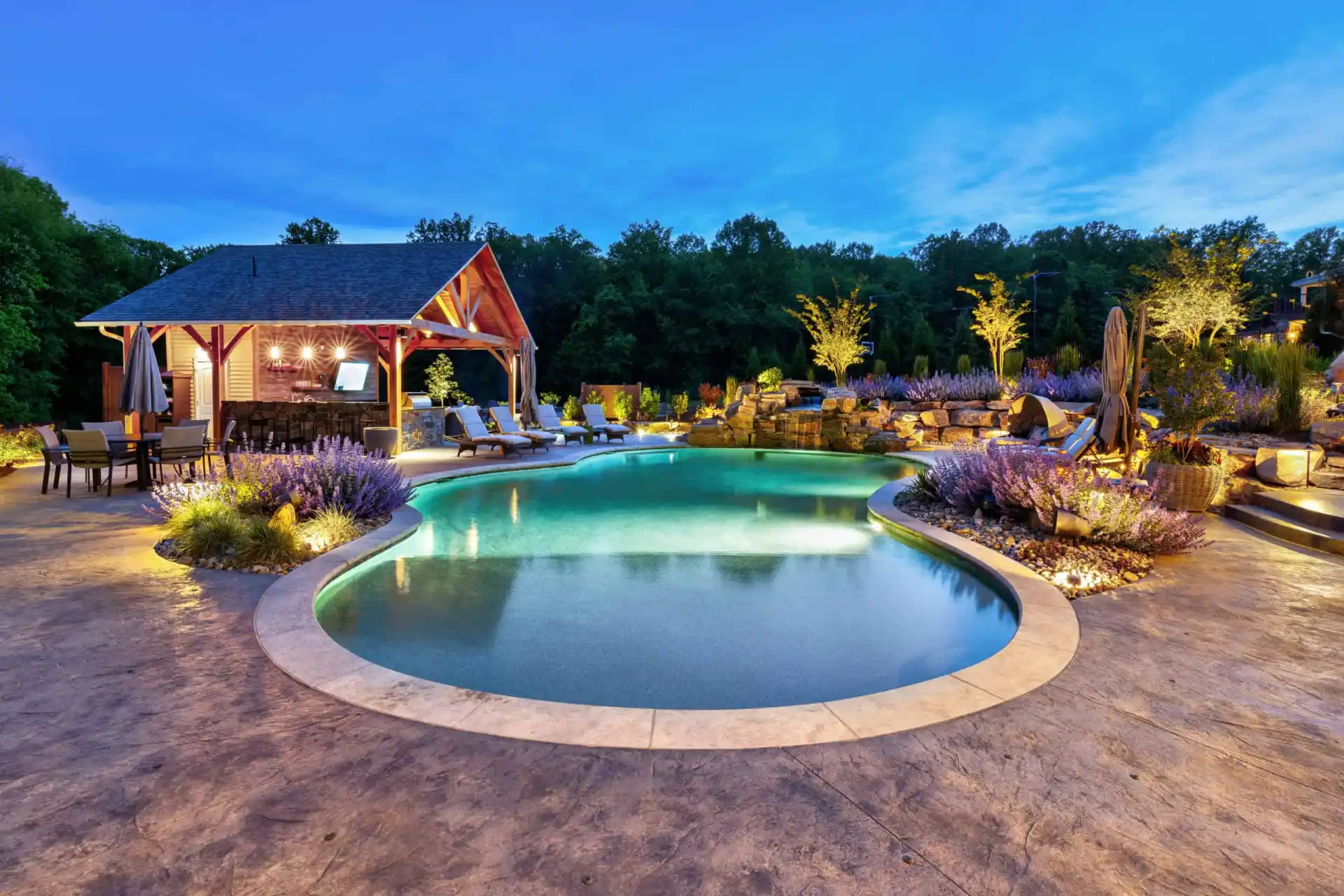 A beautifully landscaped backyard features a glowing swimming pool surrounded by flowers, lounge chairs, and a pool house at dusk, with trees and blue sky in the background.