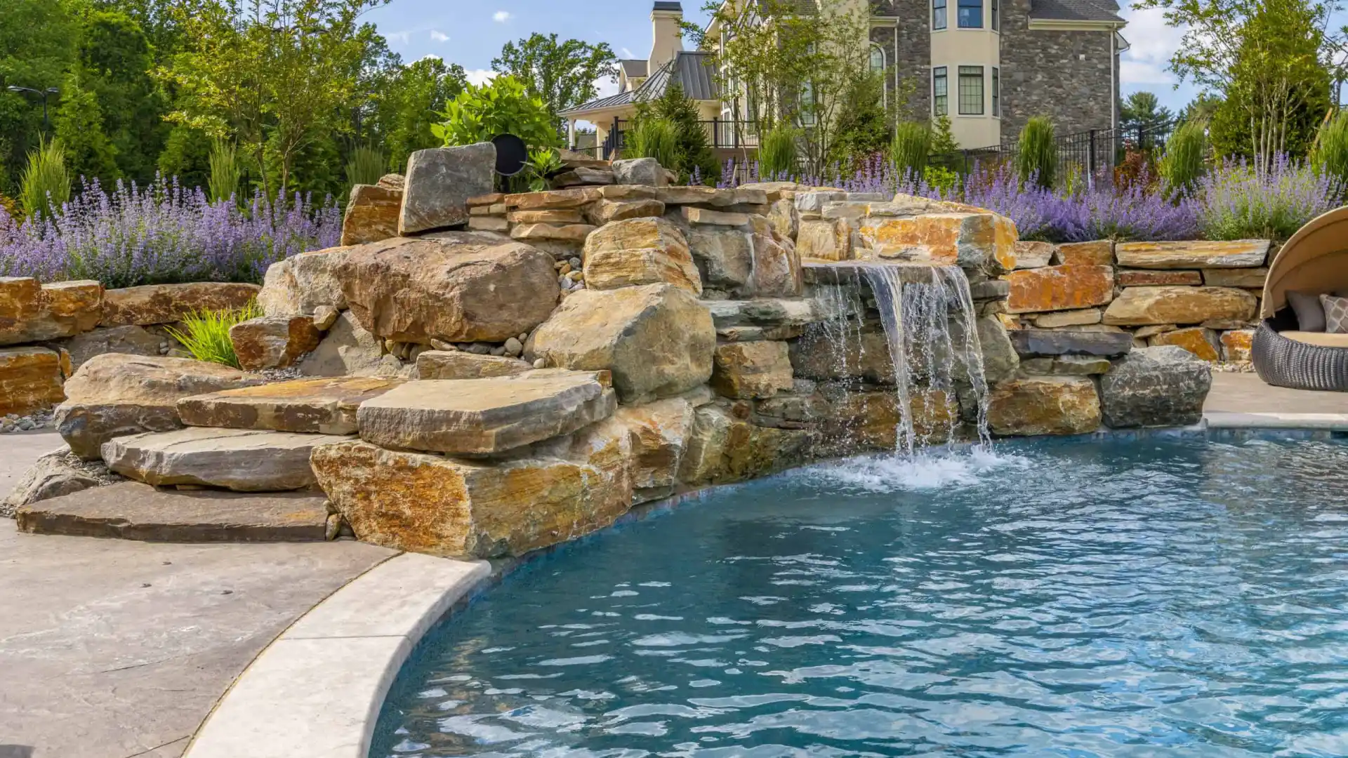 Two wooden lounge chairs face a pristine, rectangular swimming pool under a partly cloudy blue sky. The pool is surrounded by a stone patio and lush greenery, showcasing the finest hardscaping services Delaware County PA has to offer for an idyllic outdoor living experience.