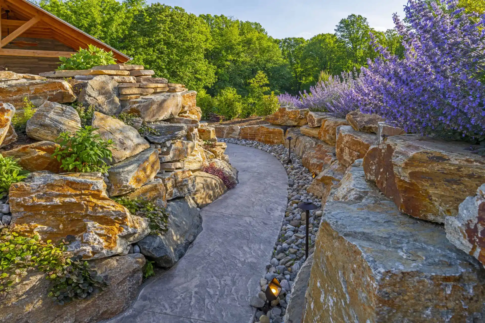 A curved stone pathway bordered by large, rust-colored rocks and vibrant purple flowering plants leads through a lush, green garden with trees in the background.