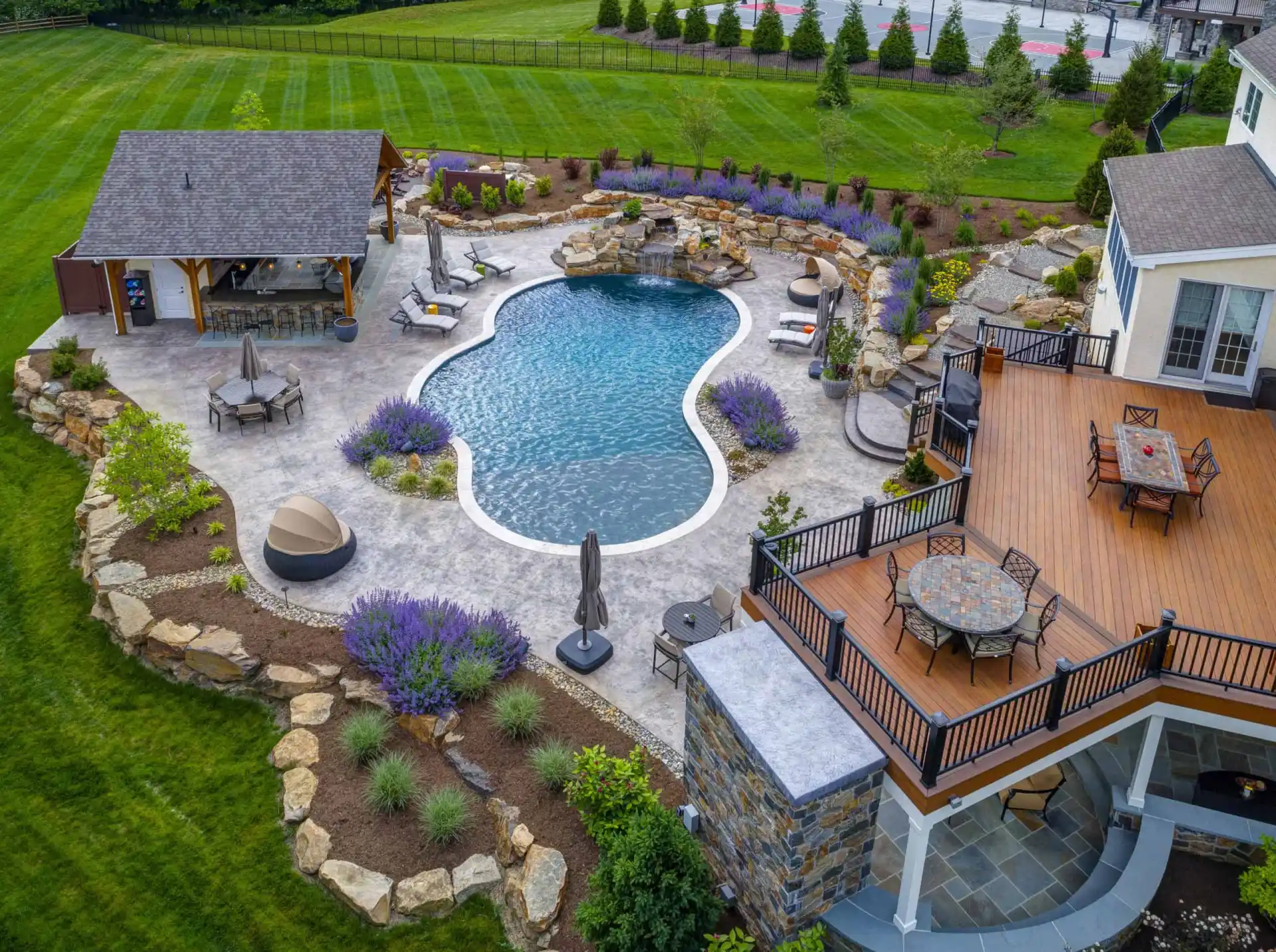 Aerial view of a backyard with a kidney-shaped pool, stone patio, lounge chairs, pool house, landscaped gardens, and a large raised deck with outdoor dining furniture.