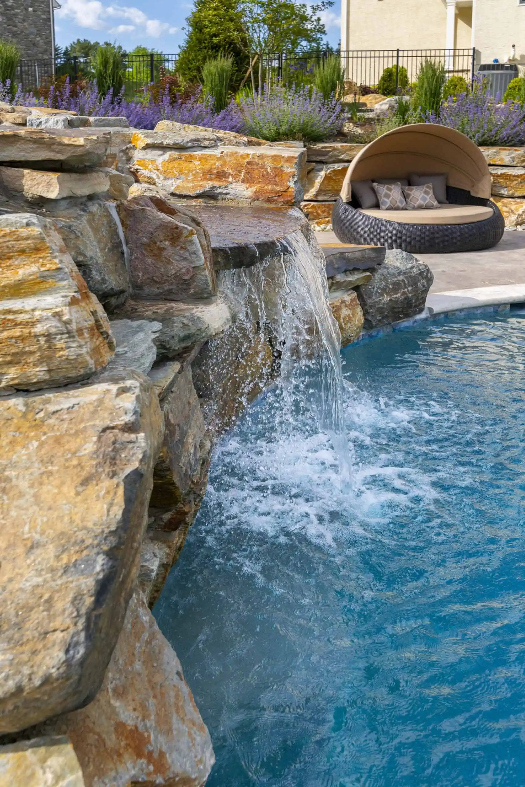 A stone waterfall flows into a bright blue swimming pool, with a round outdoor lounge chair and pillows nearby. Purple flowers and green plants line the edge of the landscaped pool area.