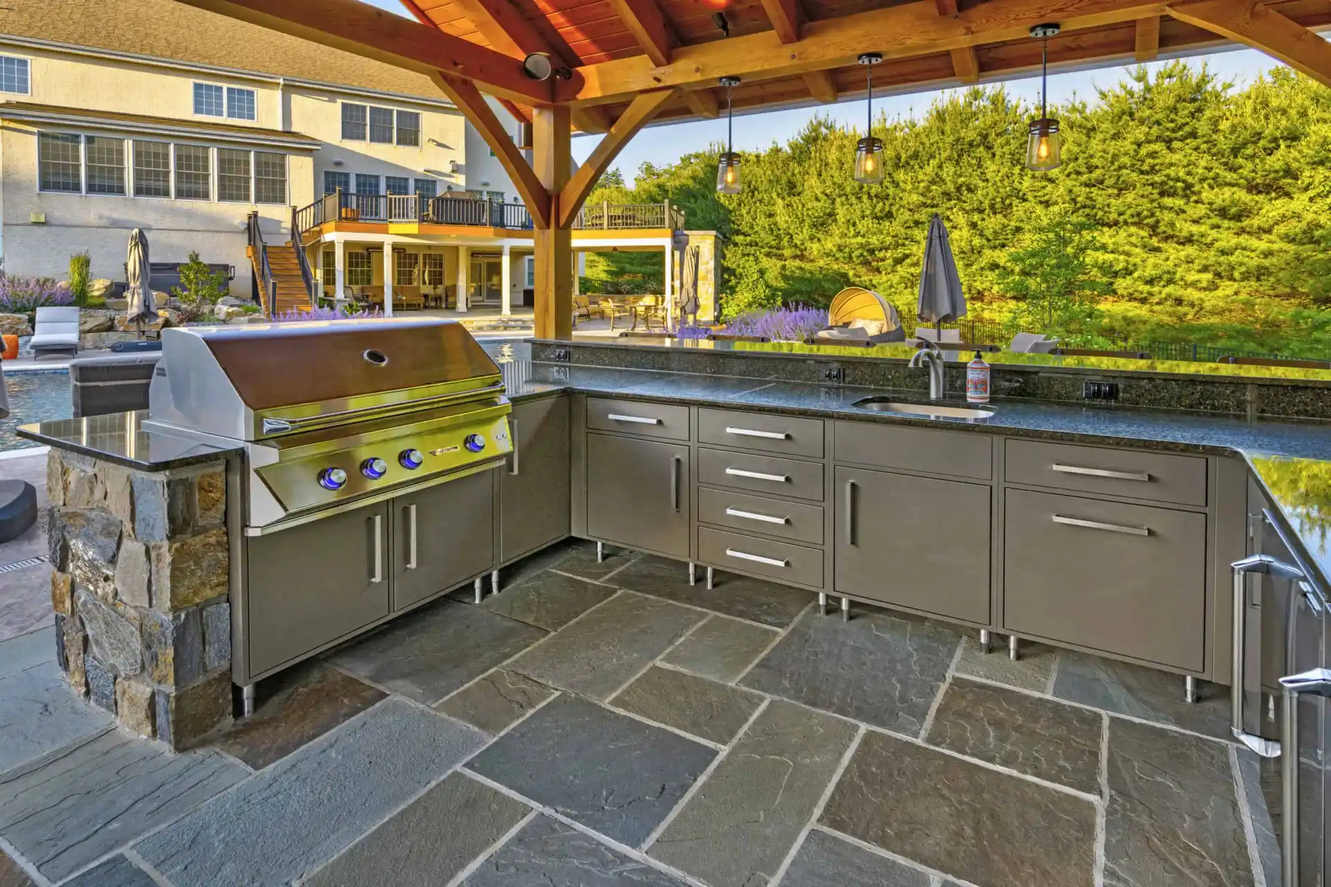 Modern outdoor kitchen under a wooden pergola with a stainless steel grill, gray cabinets, countertop, sink, and stone tile flooring, overlooking a backyard with a pool and greenery.