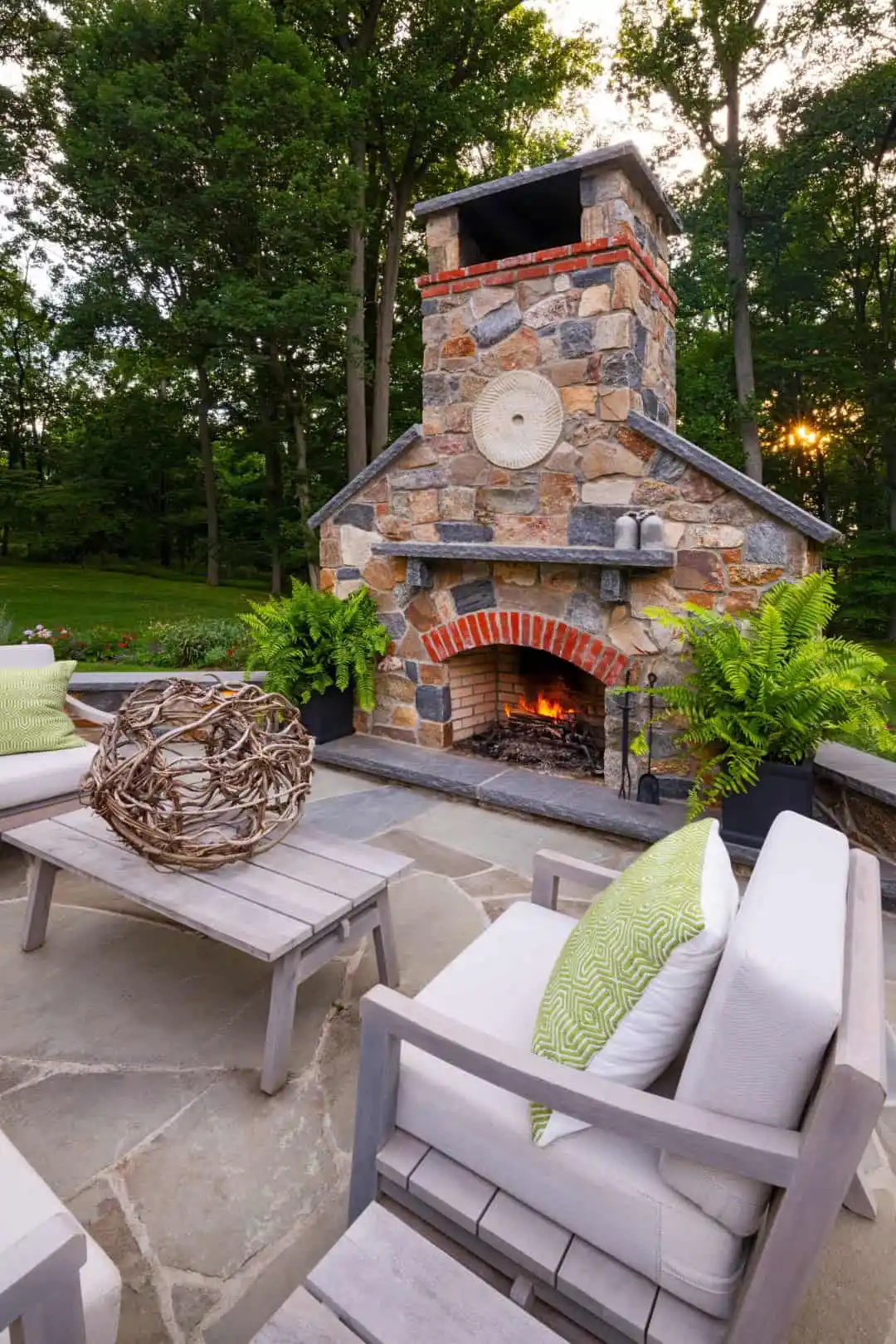 Experience elevated outdoor living in Delaware County, PA, with a stone fireplace surrounded by lush greenery. Comfy chairs and green pillows create a cozy ambiance. A decorative wicker ball graces the wooden coffee table, making this an ideal retreat enhanced by expert landscaping services.