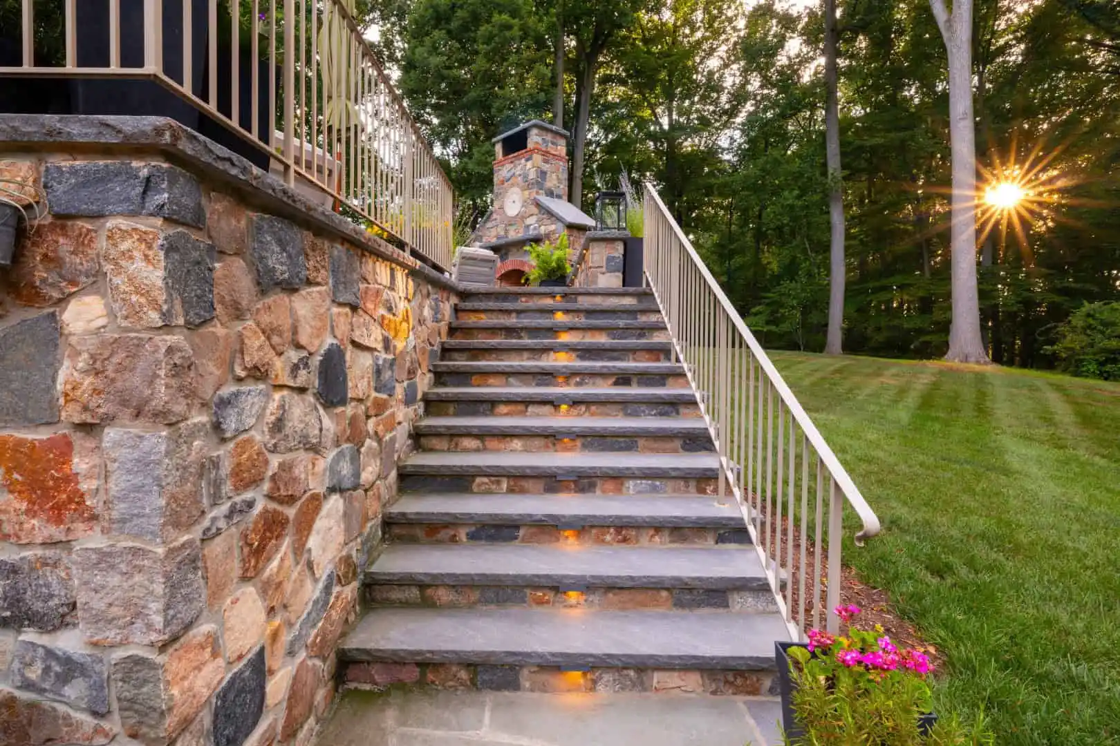 Stone steps with embedded lights lead up to a terrace featuring a stone fireplace, showcasing exquisite outdoor living in Delaware County, PA. A stone wall and metal railing line the steps as the sun peeks through trees, illuminating the grass and a vibrant flowerbed nearby.