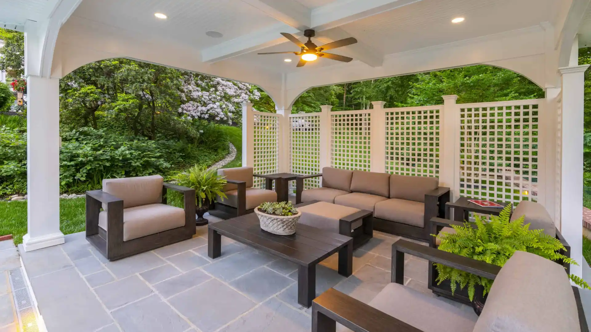 A stylish outdoor patio, perfect for enhancing your outdoor living in Delaware County, PA, features a seating set with beige cushions around a central coffee table with a plant. Two additional armchairs complement the space, covered by lattice walls and overlooking lush garden greenery. A ceiling fan is overhead.