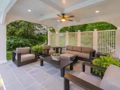 A stylish outdoor patio, perfect for enhancing your outdoor living in Delaware County, PA, features a seating set with beige cushions around a central coffee table with a plant. Two additional armchairs complement the space, covered by lattice walls and overlooking lush garden greenery. A ceiling fan is overhead.