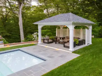 Nestled in a serene setting, this outdoor living space in Delaware County, PA, features a pool with a stone-tiled deck next to a cozy gazebo. Cushioned chairs and a table invite relaxation amid lush greenery and trees, showcasing skilled hardscaping services.
