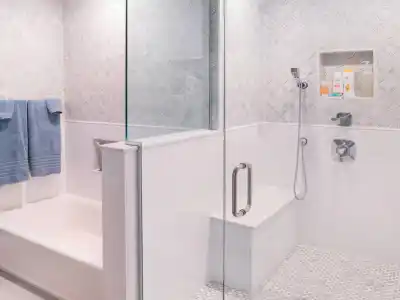 A modern bathroom features a walk-in glass shower with a built-in bench and rainfall showerhead. The walls have a textured design, and the floor has a small hexagonal tile pattern. With its seamless blend of indoor elegance and outdoor living, this space feels like an oasis.