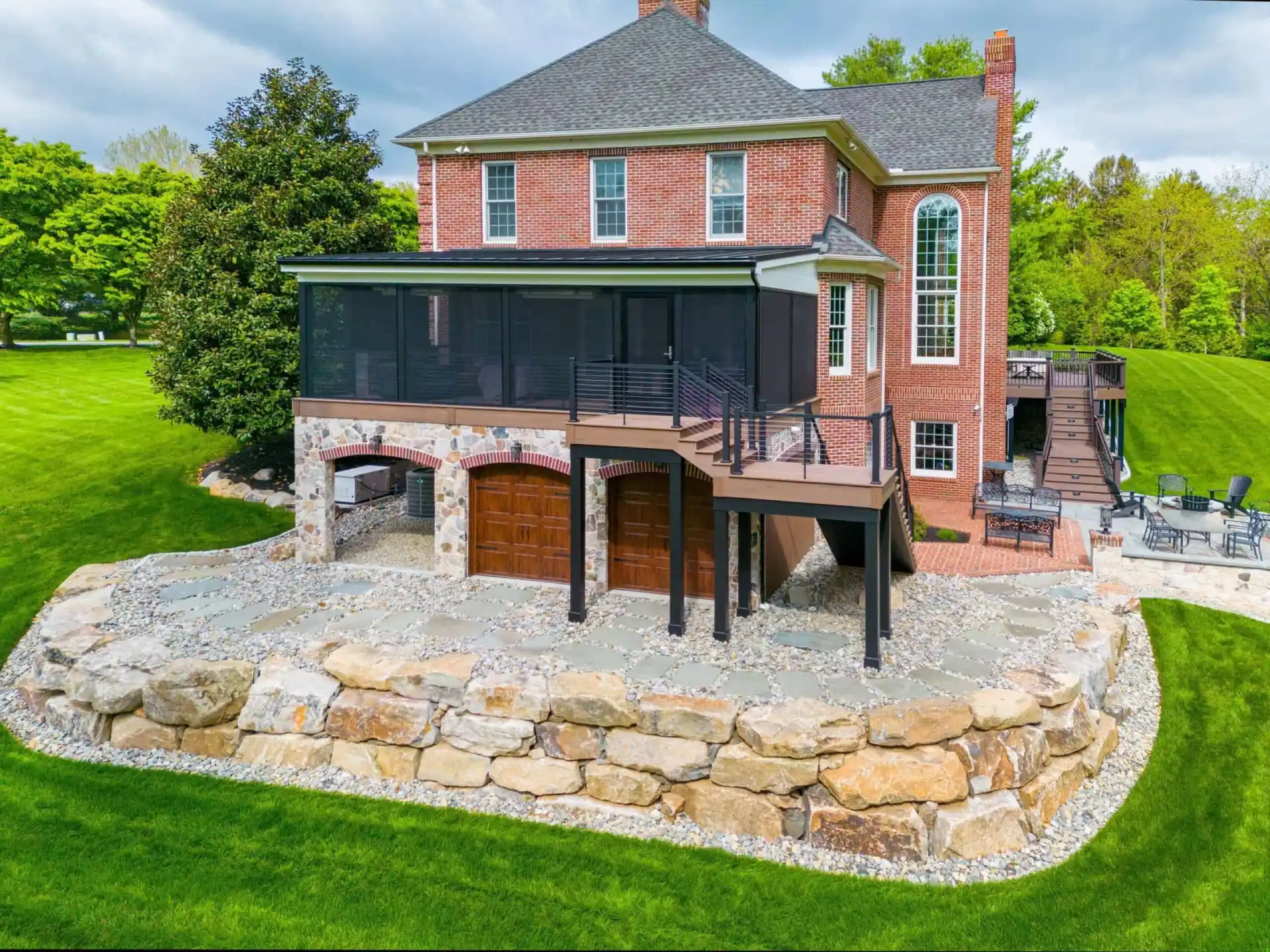 A large brick house with a stone foundation, screened porch, and multiple levels features hardscaping services in Delaware County, PA. Enjoy the wooden staircase, spacious patio, and stone-bordered lawn amidst lush grass and surrounding trees under a cloudy sky.