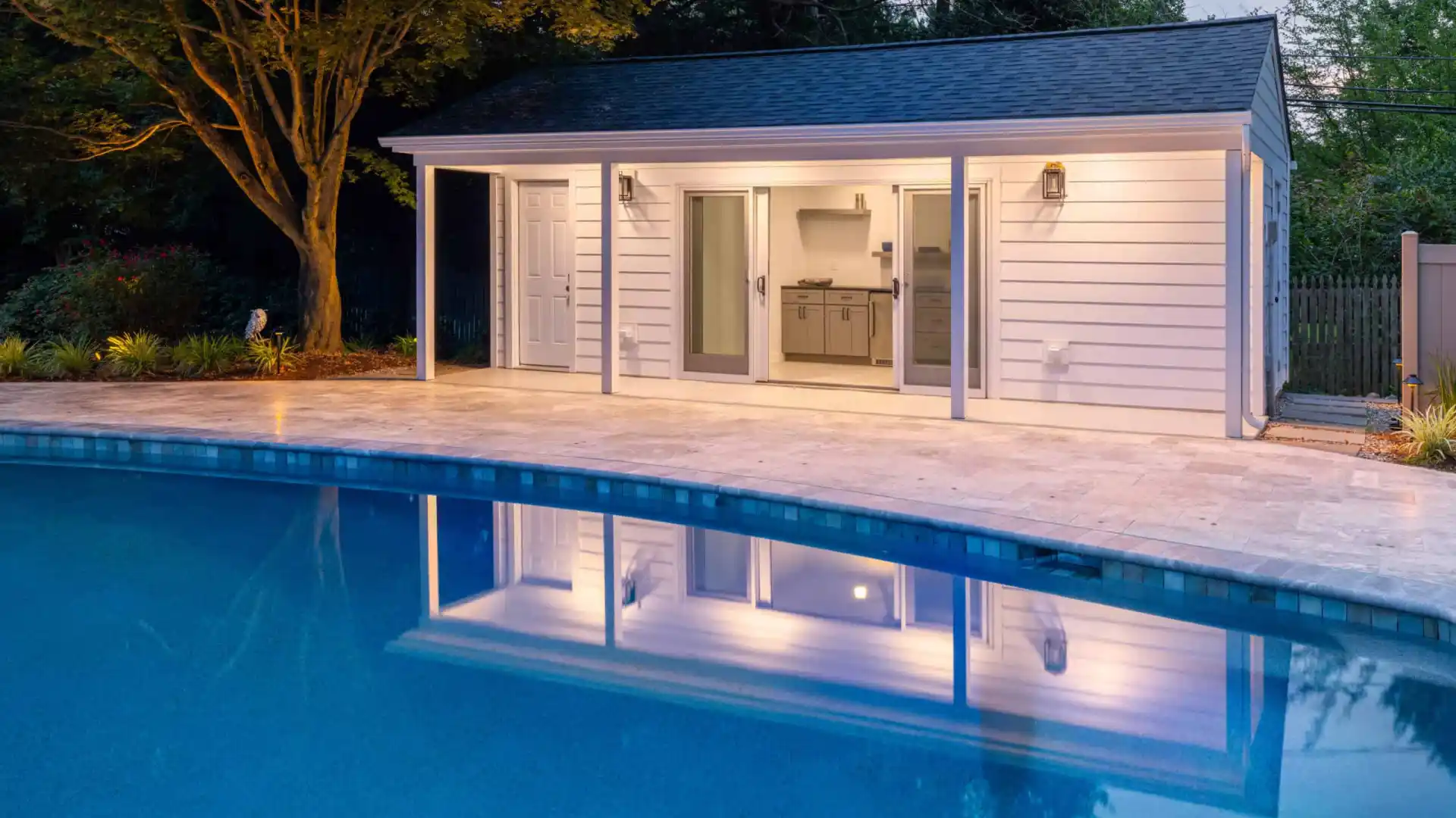 A modern pool house with white siding and large glass doors is illuminated at dusk, epitomizing outdoor living in Delaware County, PA. It stands beside a tranquil pool with reflective water, bordered by expertly crafted stone paving. Tall trees and a fence are visible in the background.