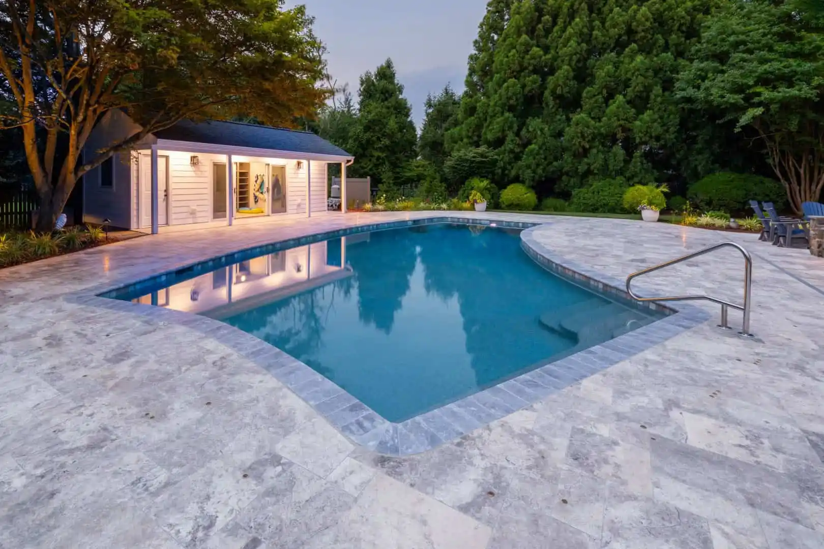 A backyard scene featuring a rectangular swimming pool with a stone deck, expertly crafted by landscaping services in Delaware County, PA. It's surrounded by lush greenery and trees, while a small pool house with large windows glows softly in the background under gentle exterior lights.