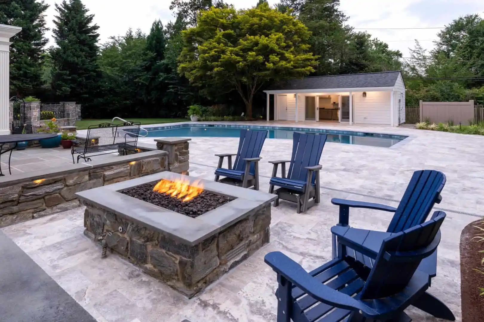 Outdoor living in Delaware County, PA, reaches new heights with a stone fire pit surrounded by blue Adirondack chairs. In the background, a pool and a cozy pool house enhance this serene backyard setting, framed perfectly by lush green trees and shrubs.