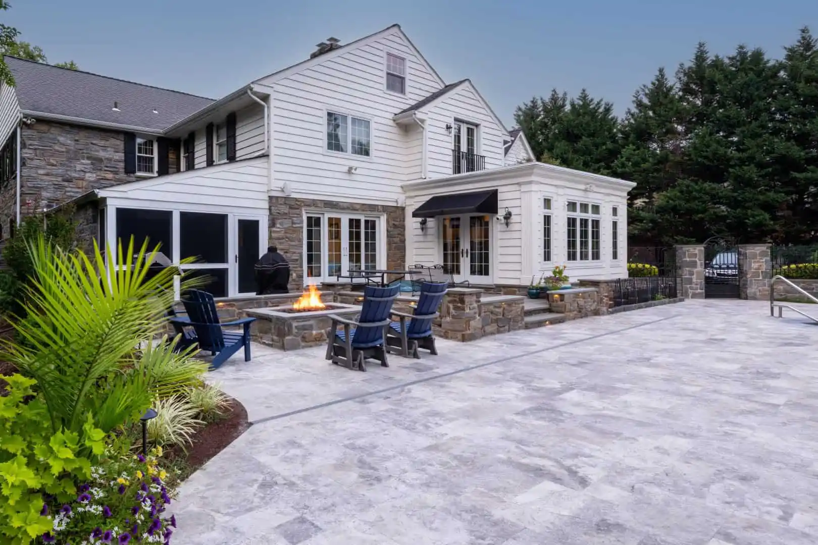 A large house with white siding and stone accents features a spacious patio perfect for outdoor living in Delaware County, PA. Blue chairs surround a fire pit amid greenery in planters, under a clear sky, creating a peaceful atmosphere enhanced by expert landscaping services.