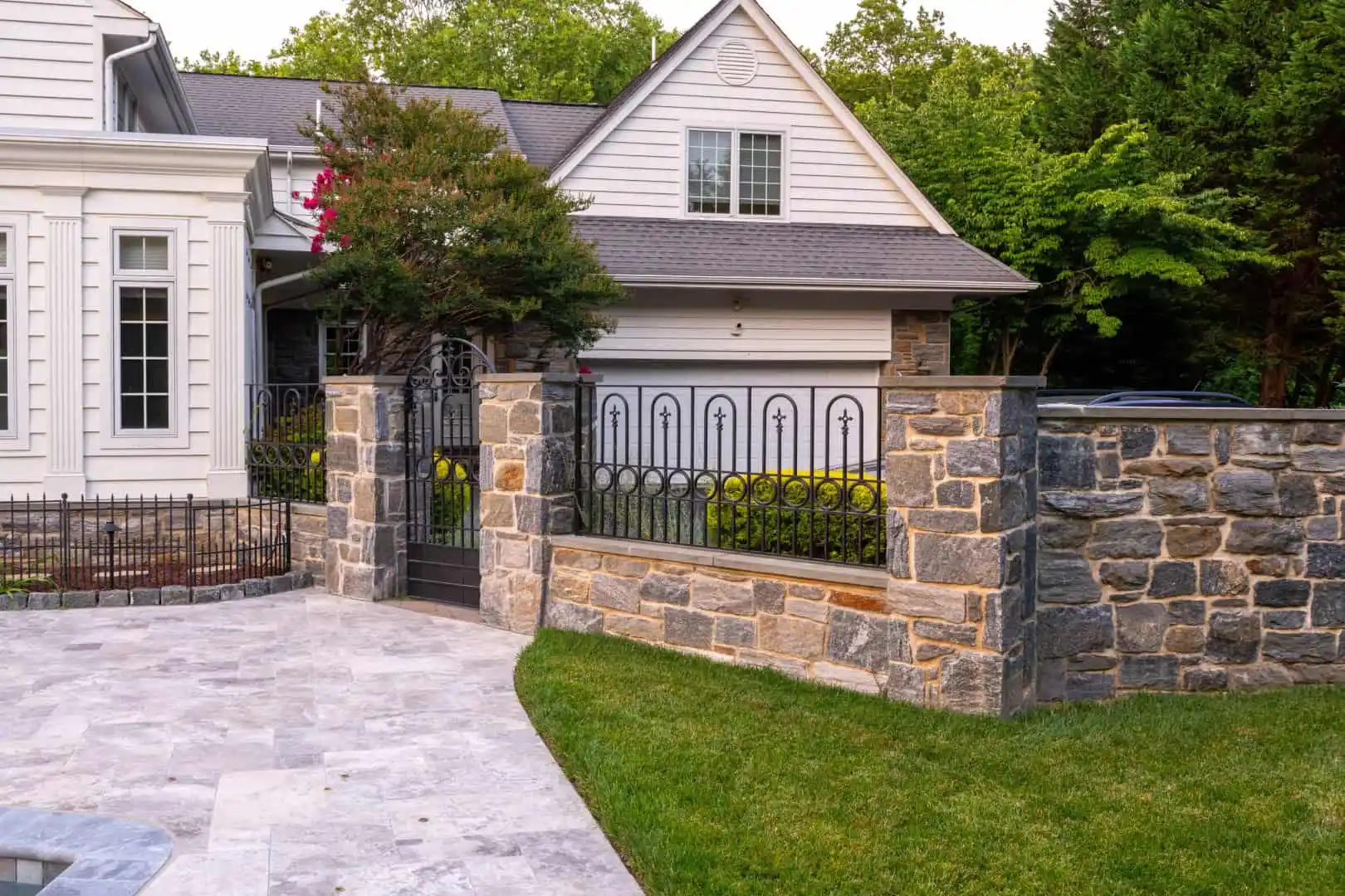 A suburban home with a gray shingled roof and white exterior is enhanced by expert hardscaping services in Delaware County, PA. A stone fence with iron gates frames the yard, featuring a tree and a patio with stone tiles, set against a lush, green lawn on the side.