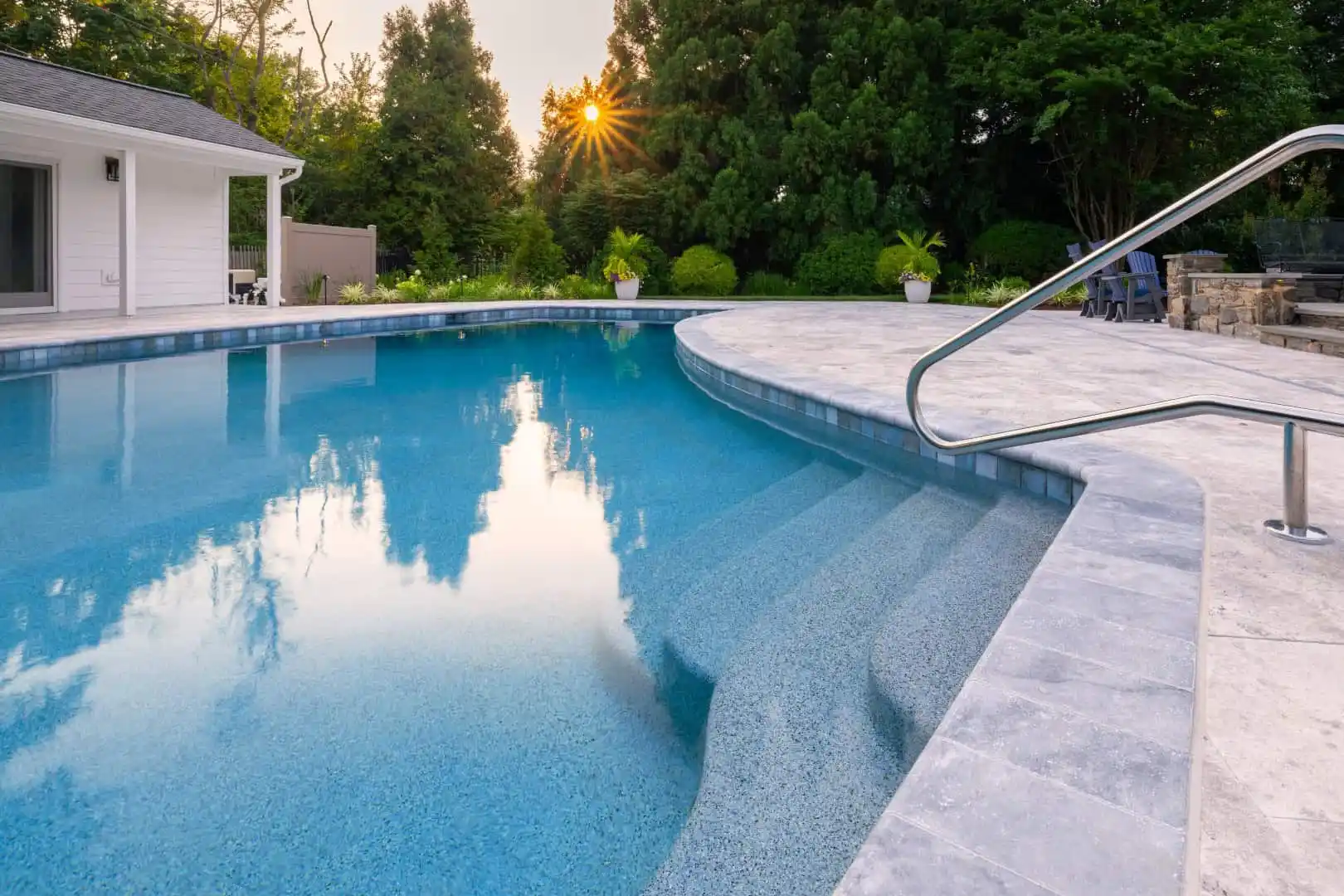 Outdoor swimming pool with crystal-clear water, nestled amid expertly crafted hardscaping. A stone deck and metal handrail complement the modern white building and lush green trees, as sunlight dances through. Two potted plants enhance the serene outdoor living in Delaware County, PA.