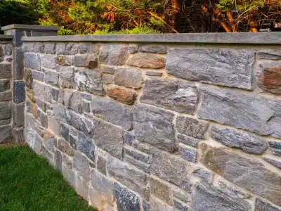 Sunlight casts its glow on a stone wall of gray and tan, arranged in a brick pattern. This picturesque scene, enhanced by professional hardscaping services in Delaware County, PA, features smooth-topped stones bordered by lush grass and sunlit leaves dancing above.