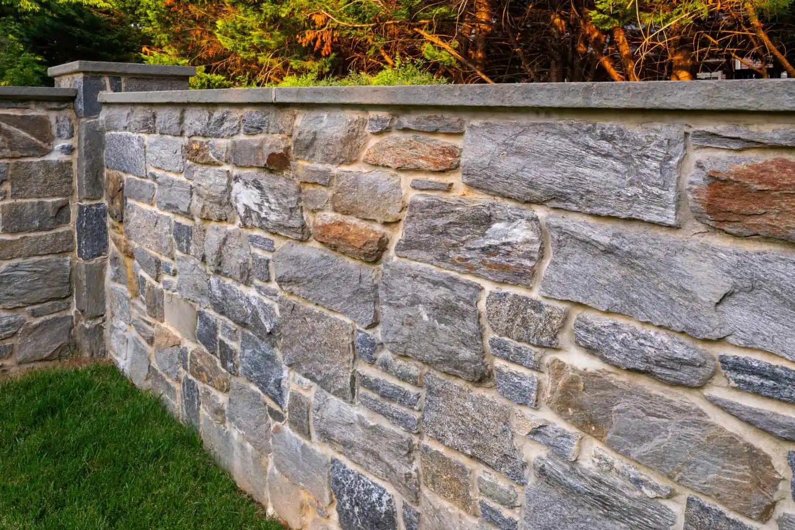 Sunlight casts its glow on a stone wall of gray and tan, arranged in a brick pattern. This picturesque scene, enhanced by professional hardscaping services in Delaware County, PA, features smooth-topped stones bordered by lush grass and sunlit leaves dancing above.