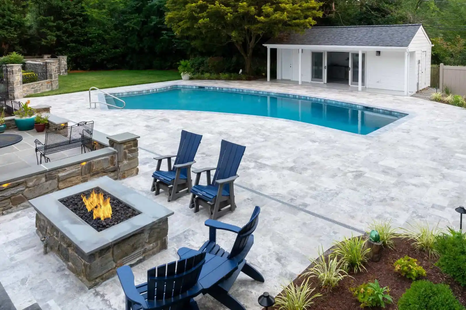 A modern backyard featuring a large swimming pool, gray stone patio, and fire pit encircled by four blue Adirondack chairs. A small white pool house stands amidst lush green landscaping, all beautifully crafted with expert hardscaping services in Delaware County, PA.