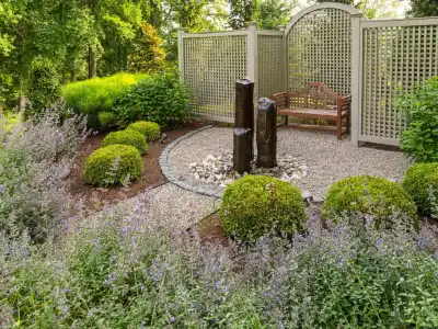 A tranquil garden features a gravel area with three vertical stone fountains, surrounded by neatly trimmed bushes, flowering plants, a wooden bench, and lattice privacy screens, all set in lush greenery.