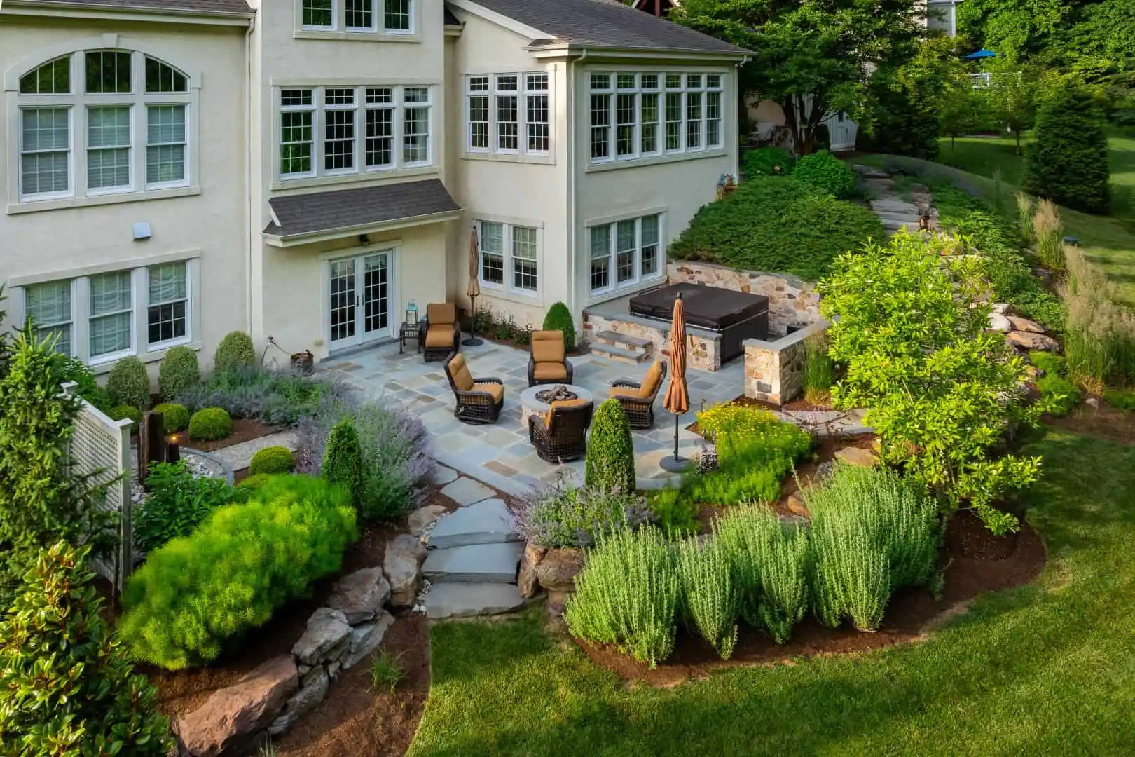 Nestled in Delaware County, PA, this elegant backyard showcases the best of outdoor living with a stone patio, comfortable furniture, and a soothing hot tub. The lush landscaping complements the two-story house's numerous windows and pristine lawn, creating a serene and inviting atmosphere.