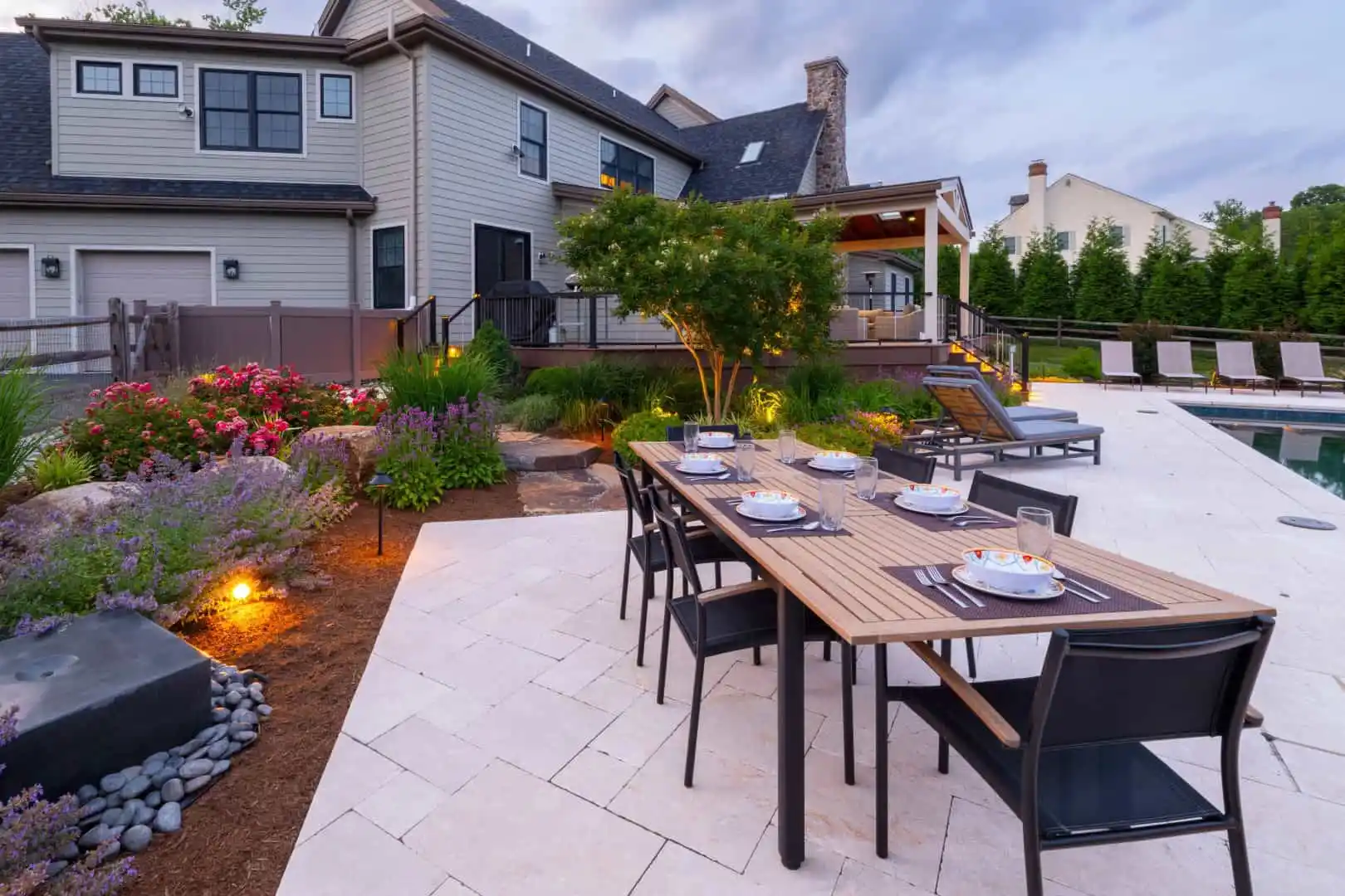 A modern backyard epitomizes outdoor living in Delaware County, PA, with a wooden dining table set on a tiled patio. Black chairs encircle it, while a landscaped garden bursting with flowers complements the pool and lounge chairs. A spacious house looms in the background under a cloudy sky.